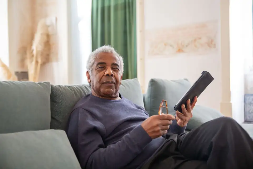 Senhor sentado no sofá segurando óculos e um tablet, analisando as modalidades de aposentadoria